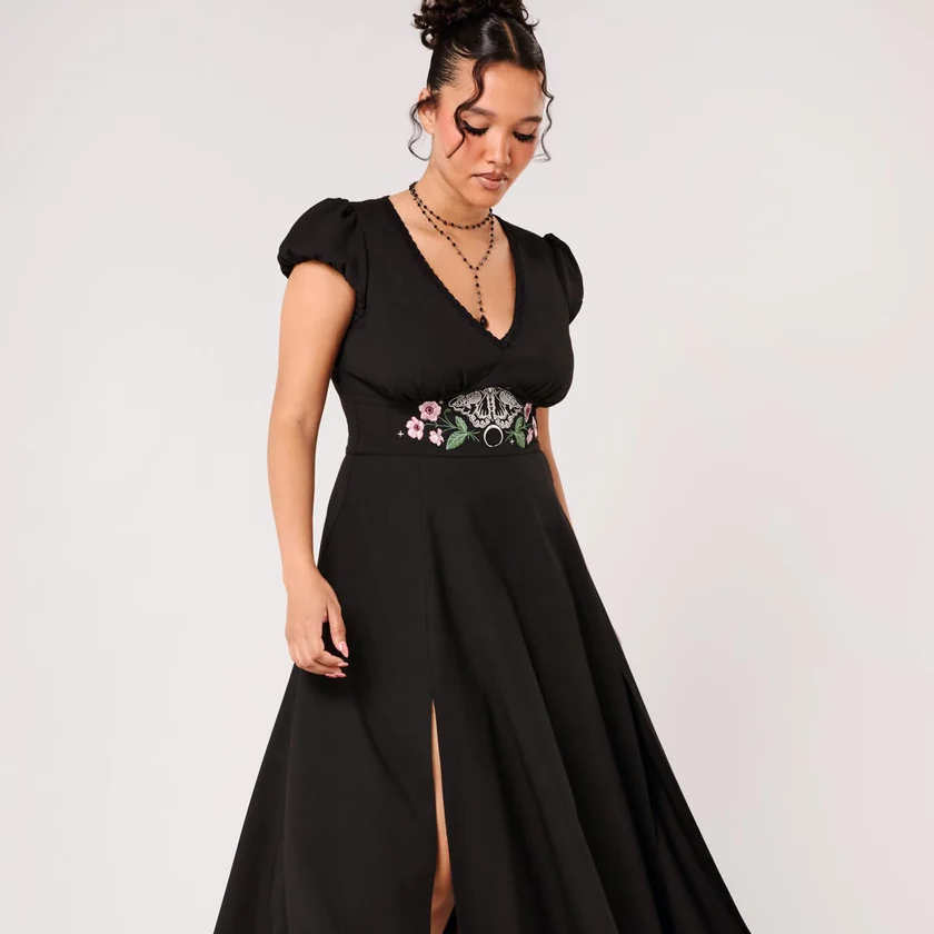 A woman modeling a black midi dress with short puff sleeves, a V-neckline, and a high thigh slit. The dress features a decorative embroidered waistband with pink, white, and green floral details.