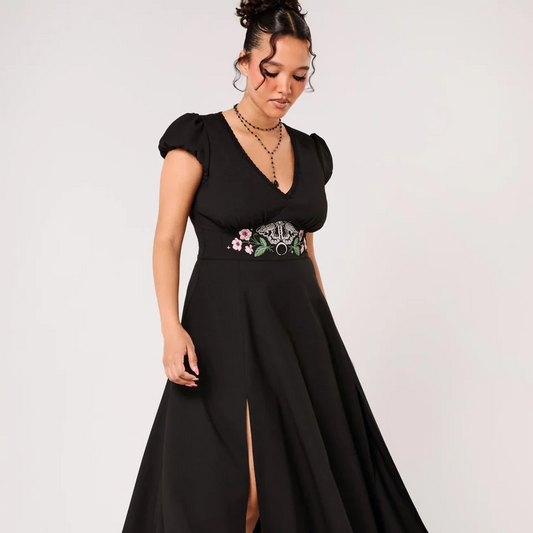 A woman modeling a black midi dress with short puff sleeves, a V-neckline, and a high thigh slit. The dress features a decorative embroidered waistband with pink, white, and green floral details.