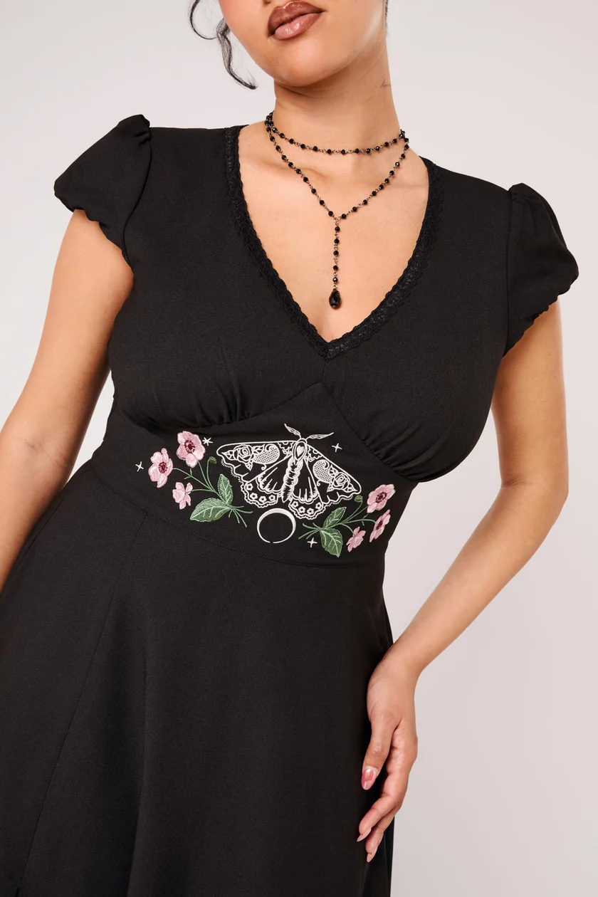 A woman wearing a black V-neck dress with short puff sleeves and a fitted waist featuring embroidered details of a moth and pink flowers, paired with black beaded necklaces against a light gray background.