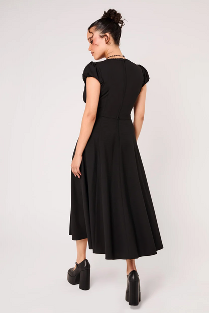 A woman modeling a black midi dress from the back, featuring short cap sleeves, a fitted waist, and a flared skirt, paired with black platform heels against a light gray studio background.