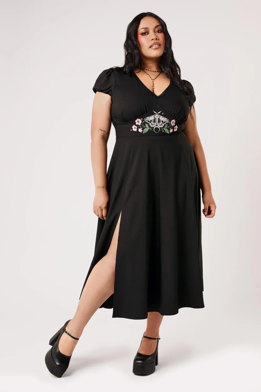 A woman modeling a black midi dress with short puff sleeves, a V-neckline, and a high side slit. The dress features a decorative embroidered waistband with green leaves and pink flowers.