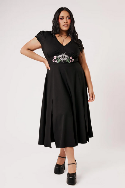 A woman modeling a black midi dress with short puff sleeves, a V-neckline, and a floral embroidered waistband, paired with black platform heels against a light gray studio background.