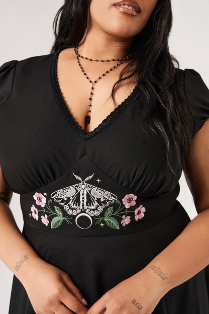A close-up of a woman wearing a black V-neck dress with lace trim, featuring a central embroidered design of a white moth, green leaves, pink flowers, and a silver ring motif.