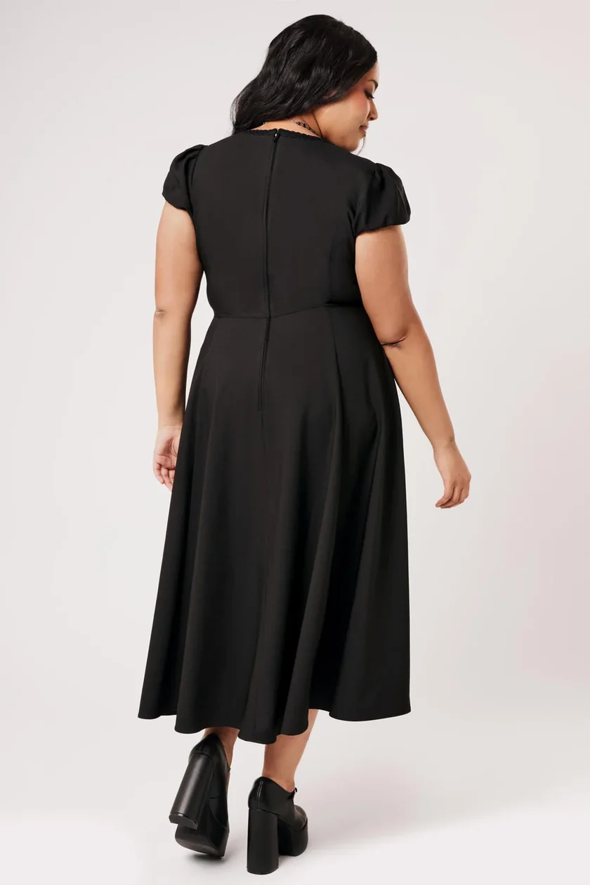 A woman shown from the back wearing a black midi dress with short puff sleeves and a flared skirt, paired with black ankle boots, against a light gray studio background.