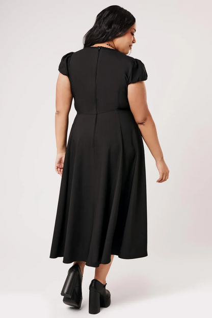 A woman shown from the back wearing a black midi dress with short puff sleeves and a flared skirt, paired with black ankle boots, against a light gray studio background.