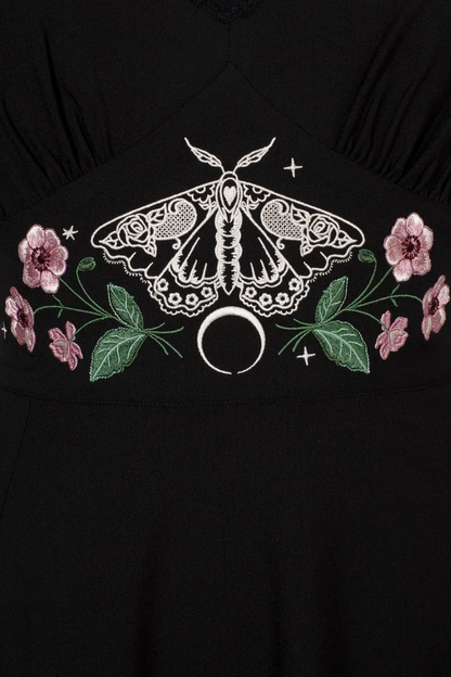 Black dress featuring a white embroidered moth and silver crescent moon motif, flanked by pink flowers and green leaves.