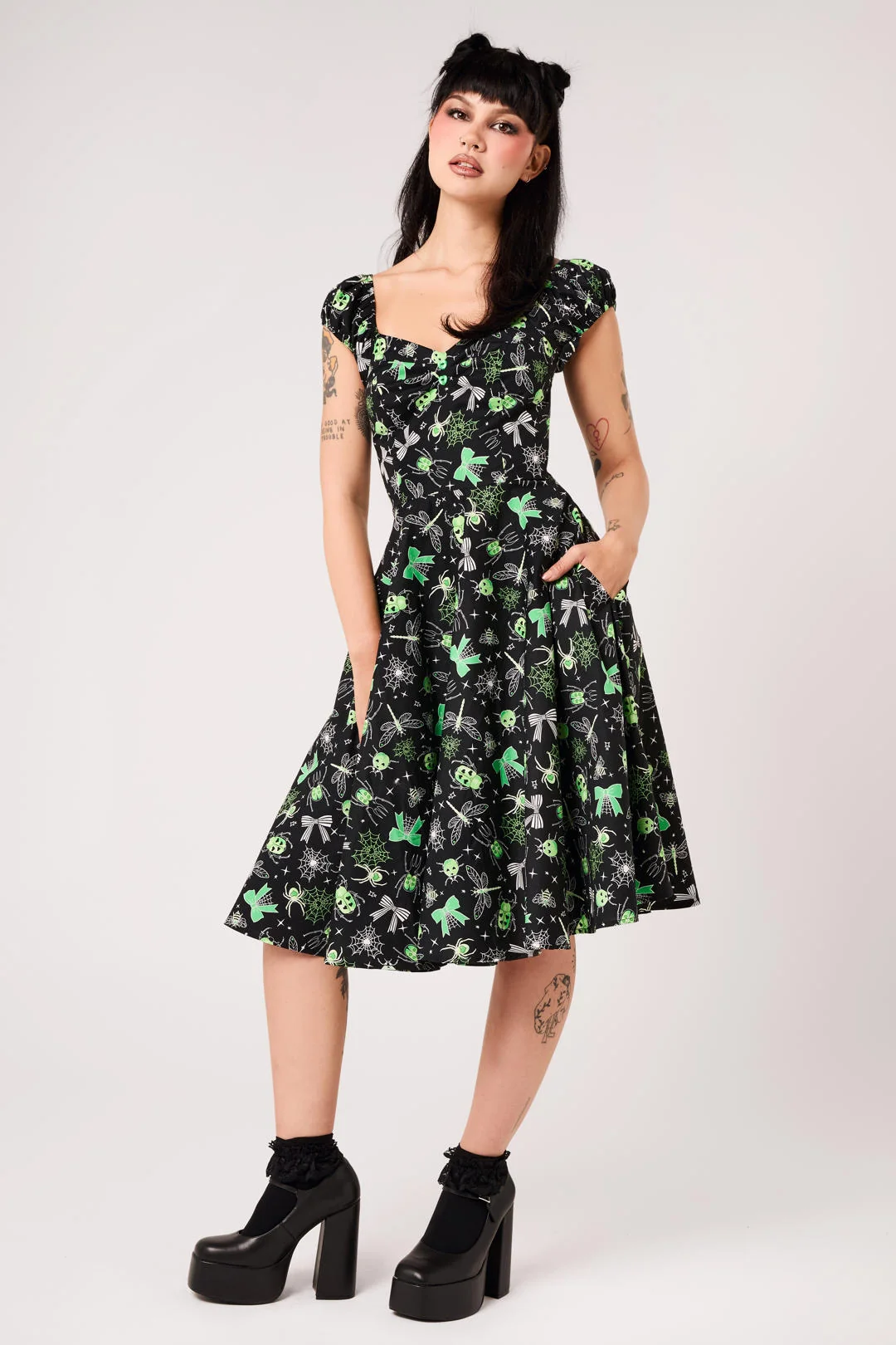A woman modeling a black fit-and-flare A-line dress with a green and gray floral pattern, featuring cap sleeves, a square neckline, and side pockets. She is wearing black platform ankle boots and a black hair accessory, standing against a light gray studio background.