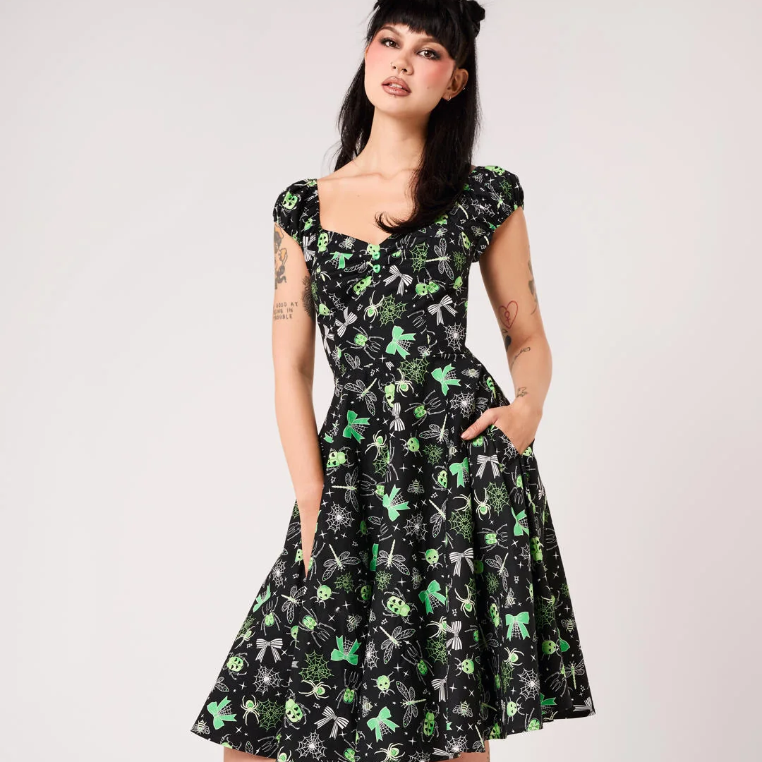 A woman modeling a black fit-and-flare A-line dress with a green and gray floral pattern, featuring cap sleeves, a square neckline, and side pockets. She is wearing black platform ankle boots and a black hair accessory, standing against a light gray studio background.