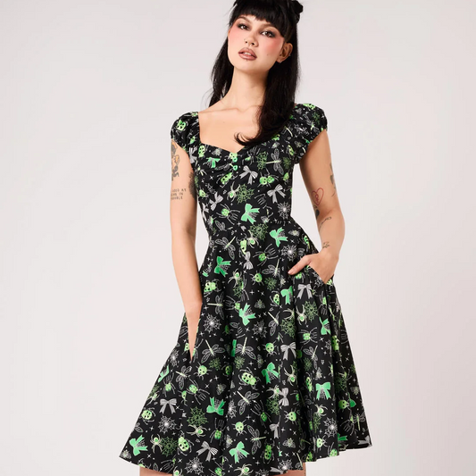 A woman modeling a black fit-and-flare A-line dress with a green and gray floral pattern, featuring cap sleeves, a square neckline, and side pockets. She is wearing black platform ankle boots and a black hair accessory, standing against a light gray studio background.