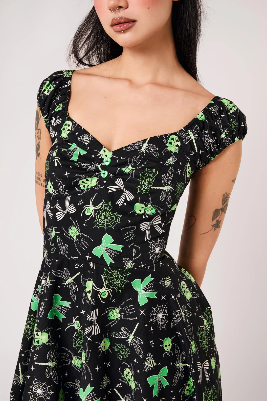A woman wearing a black off-the-shoulder dress with a green and white gothic insect pattern, including dragonflies, butterflies, and skulls, shown against a plain light gray background.