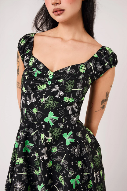 A woman wearing a black off-the-shoulder dress with a green and white gothic insect pattern, including dragonflies, butterflies, and skulls, shown against a plain light gray background.