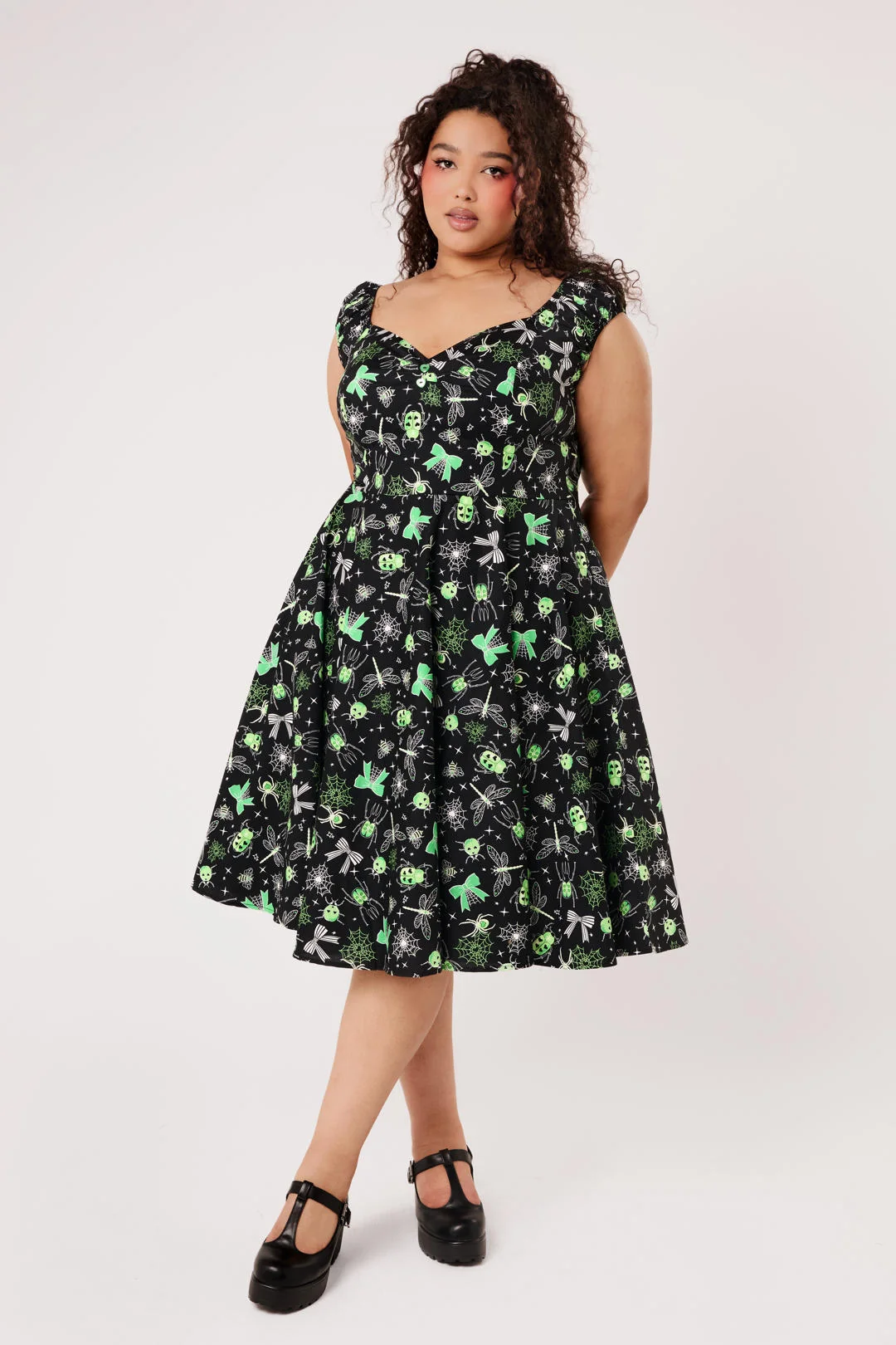 A woman modeling a sleeveless black swing dress with a green and white skull-and-crossbones print, paired with black Mary Jane shoes, against a plain light background.