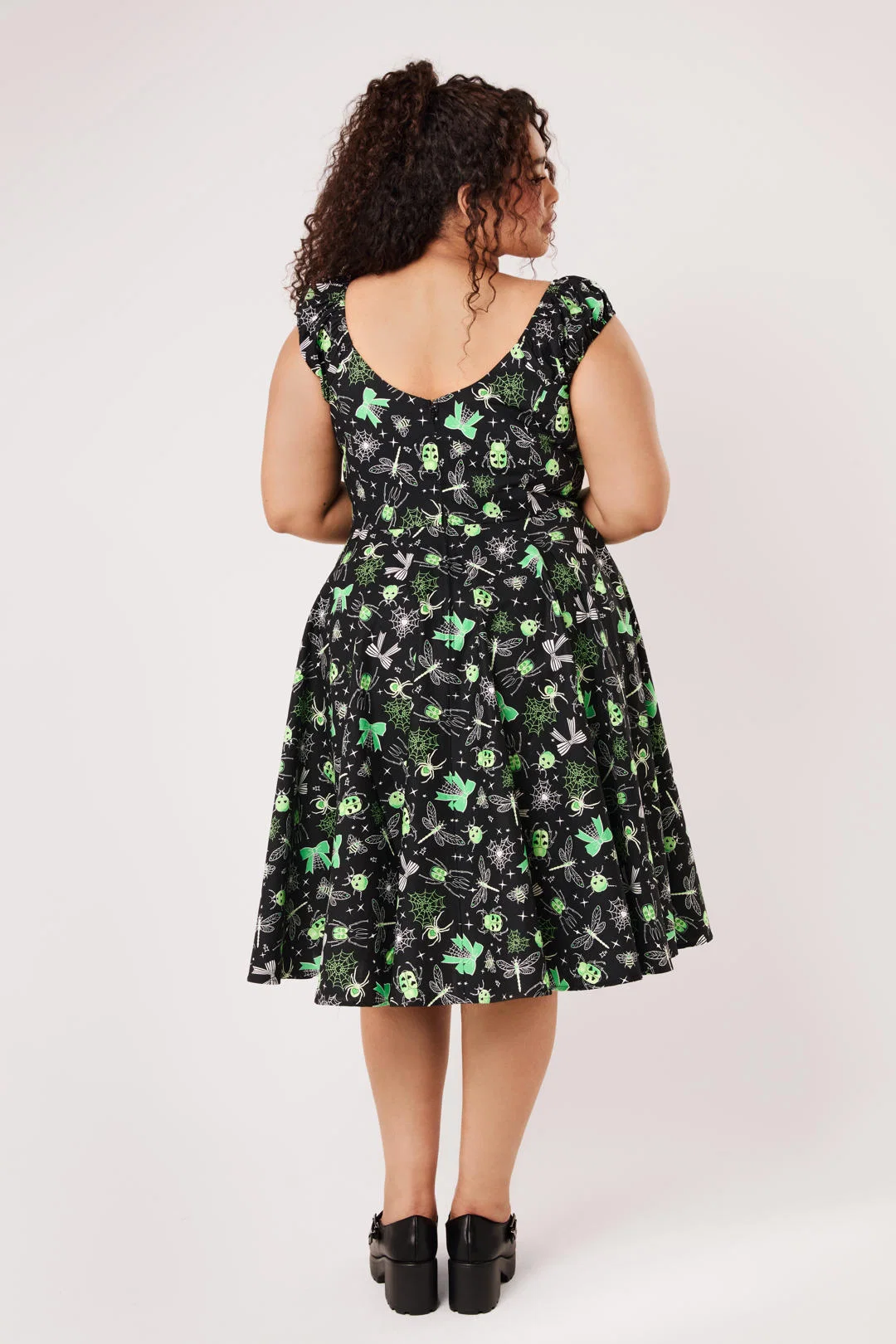 A woman viewed from the back wearing a sleeveless black dress with a green and gray floral pattern, paired with black platform shoes, against a light gray studio background.