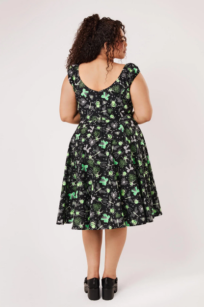 A woman viewed from the back wearing a sleeveless black dress with a green and gray floral pattern, paired with black platform shoes, against a light gray studio background.