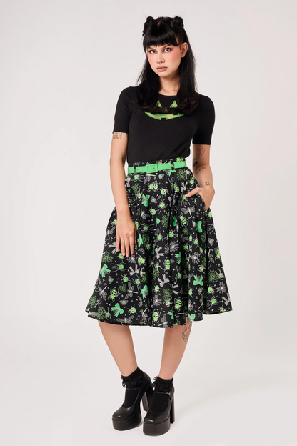A model wearing a black short-sleeve t-shirt with a green graphic print and a black pleated A-line skirt featuring a green and white skull-and-crossbones pattern, styled with a bright green belt and black platform boots against a white studio background.
