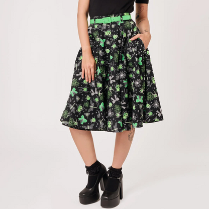 A model wearing a black short-sleeve t-shirt with a green graphic print and a black pleated A-line skirt featuring a green and white skull-and-crossbones pattern, styled with a bright green belt and black platform boots against a white studio background.
