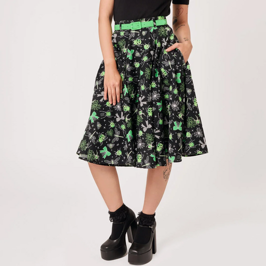 A model wearing a black short-sleeve t-shirt with a green graphic print and a black pleated A-line skirt featuring a green and white skull-and-crossbones pattern, styled with a bright green belt and black platform boots against a white studio background.