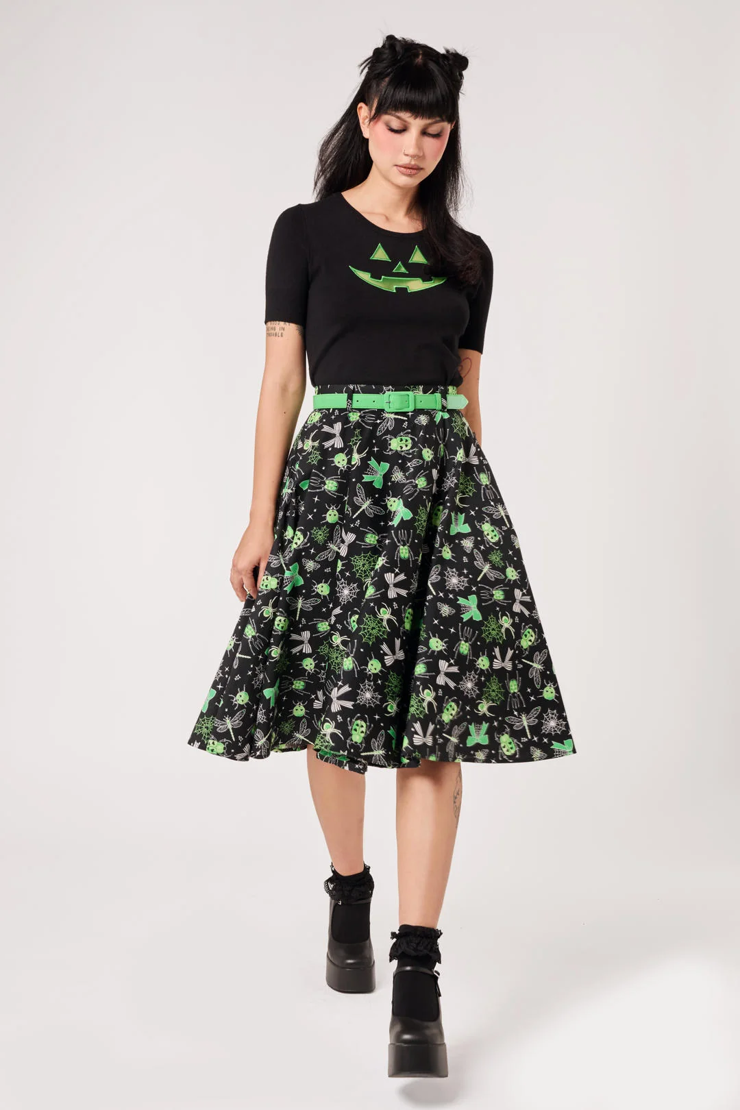 A model wearing a black short-sleeve t-shirt with a green graphic, a black midi skirt with a green and gray pattern, a bright green belt, and black platform boots, shot against a light gray studio background.