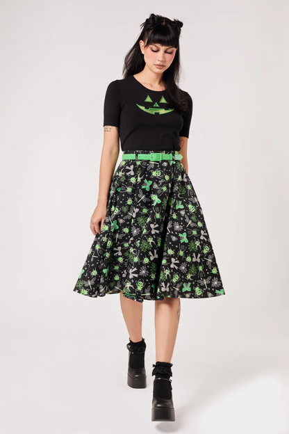 A model wearing a black short-sleeve t-shirt with a green graphic, a black midi skirt with a green and gray pattern, a bright green belt, and black platform boots, shot against a light gray studio background.