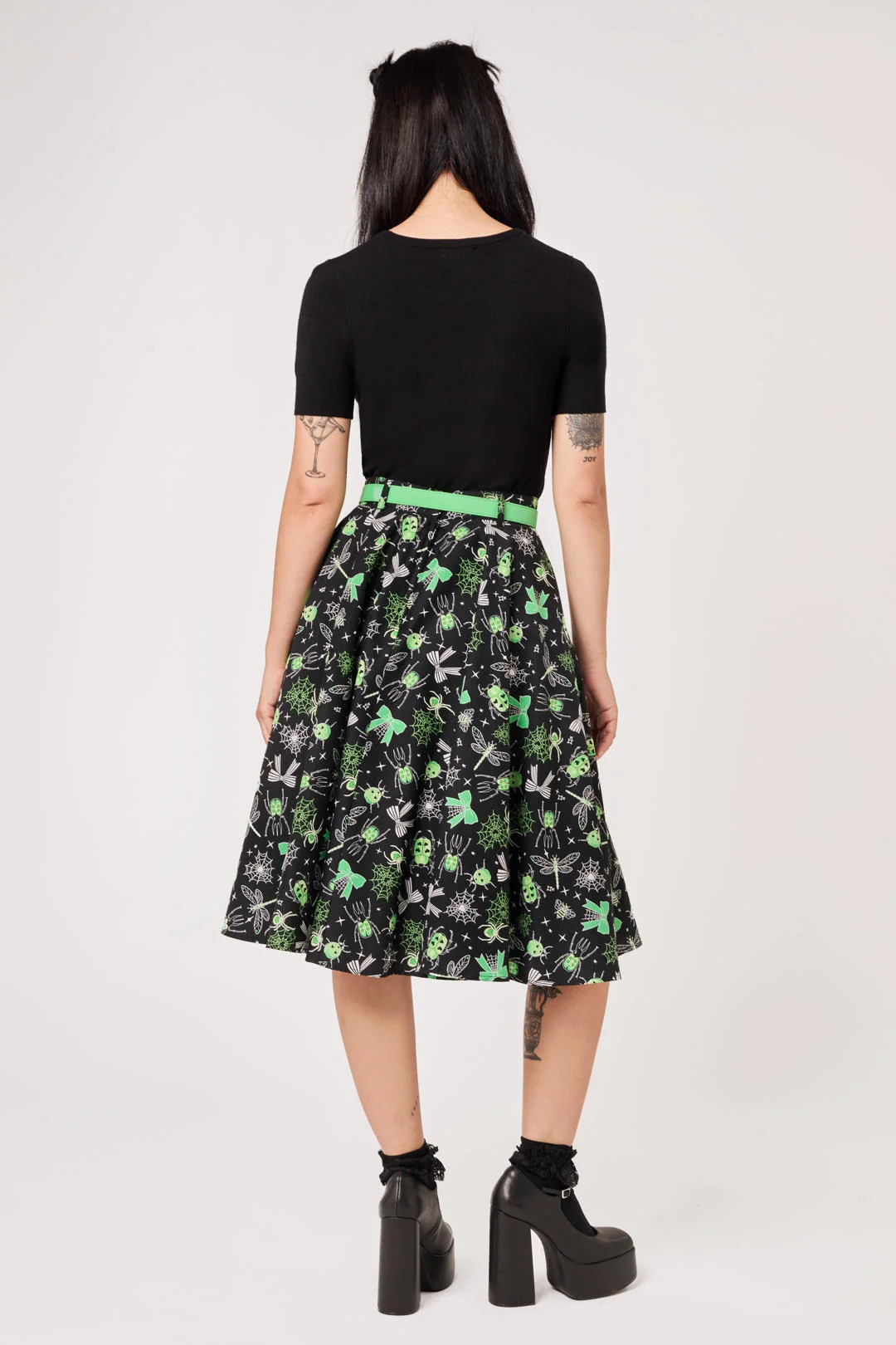 A model shown from the back wearing a black short-sleeve top and a black A-line skirt with a green and white floral pattern, paired with a bright green belt and black platform boots.