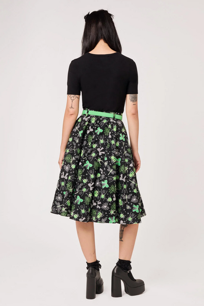 A model shown from the back wearing a black short-sleeve top and a black A-line skirt with a green and white floral pattern, paired with a bright green belt and black platform boots.