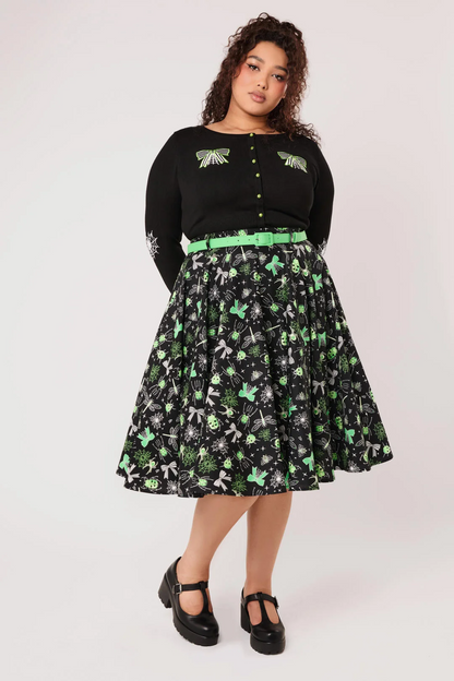 A woman modeling a black long-sleeve top with green embroidered wing details and a black midi skirt featuring a green and white dragonfly print, paired with a bright green belt and black Mary Jane shoes, against a light gray studio background.