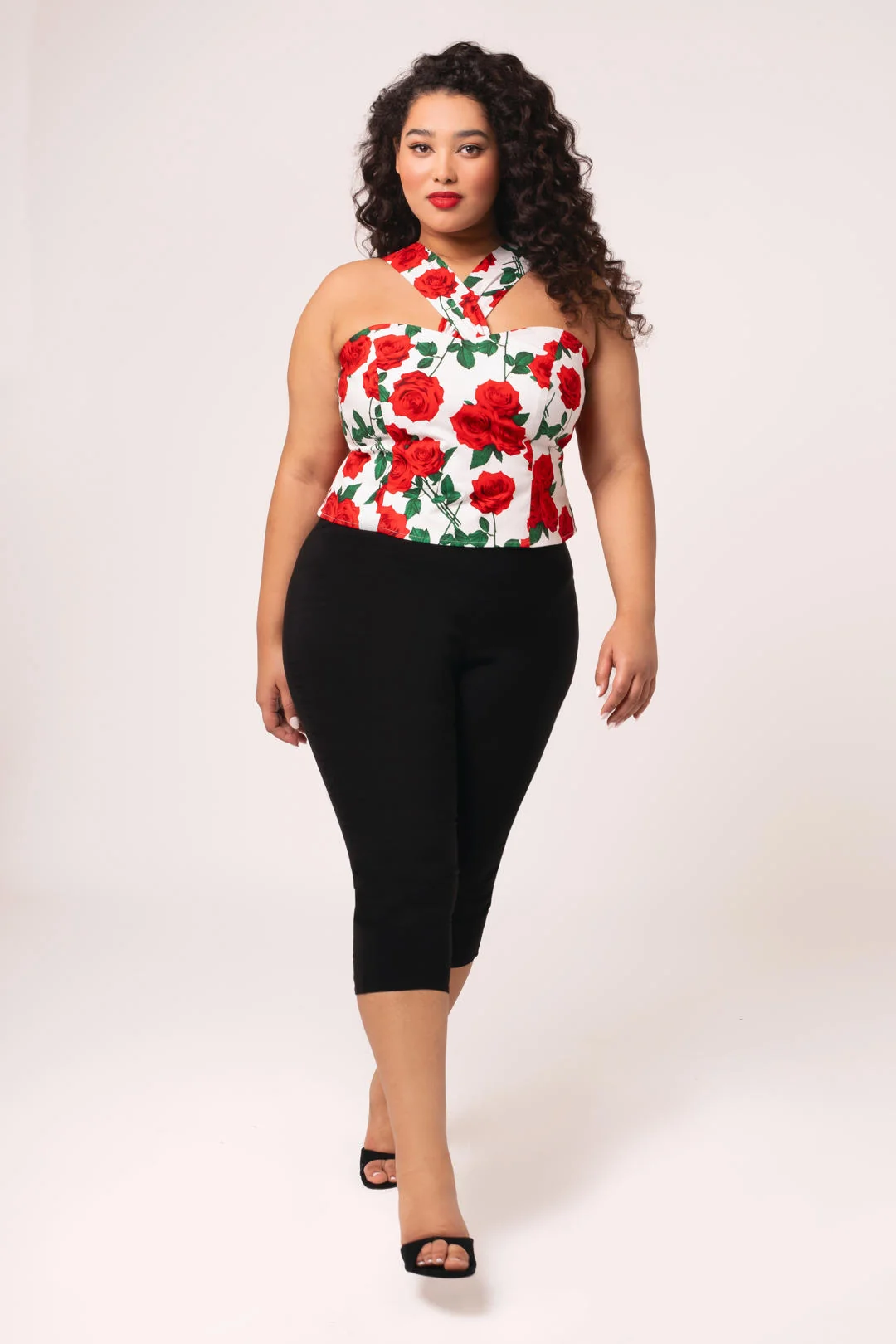 A woman modeling a halter-style top with a red rose floral print on a white background, paired with black capri pants and black sandals, against a light studio backdrop.