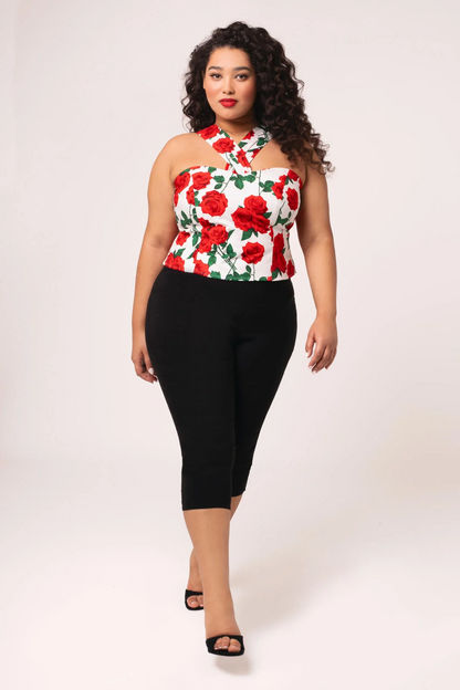 A woman modeling a halter-style top with a red rose floral print on a white background, paired with black capri pants and black sandals, against a light studio backdrop.