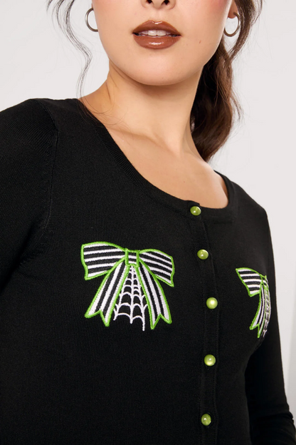 A woman modeling a black long-sleeve knit top with green and white spider-themed embroidery on the chest and sleeves, paired with black pants, against a light gray studio background.
