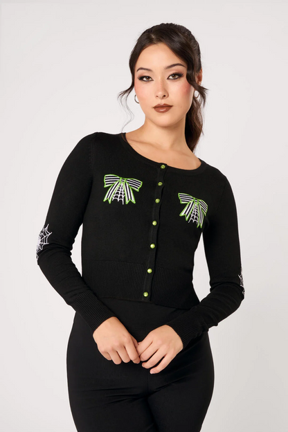 A woman modeling a black long-sleeve knit top with green and white spider-themed embroidery on the chest and sleeves, paired with black pants, against a light gray studio background.