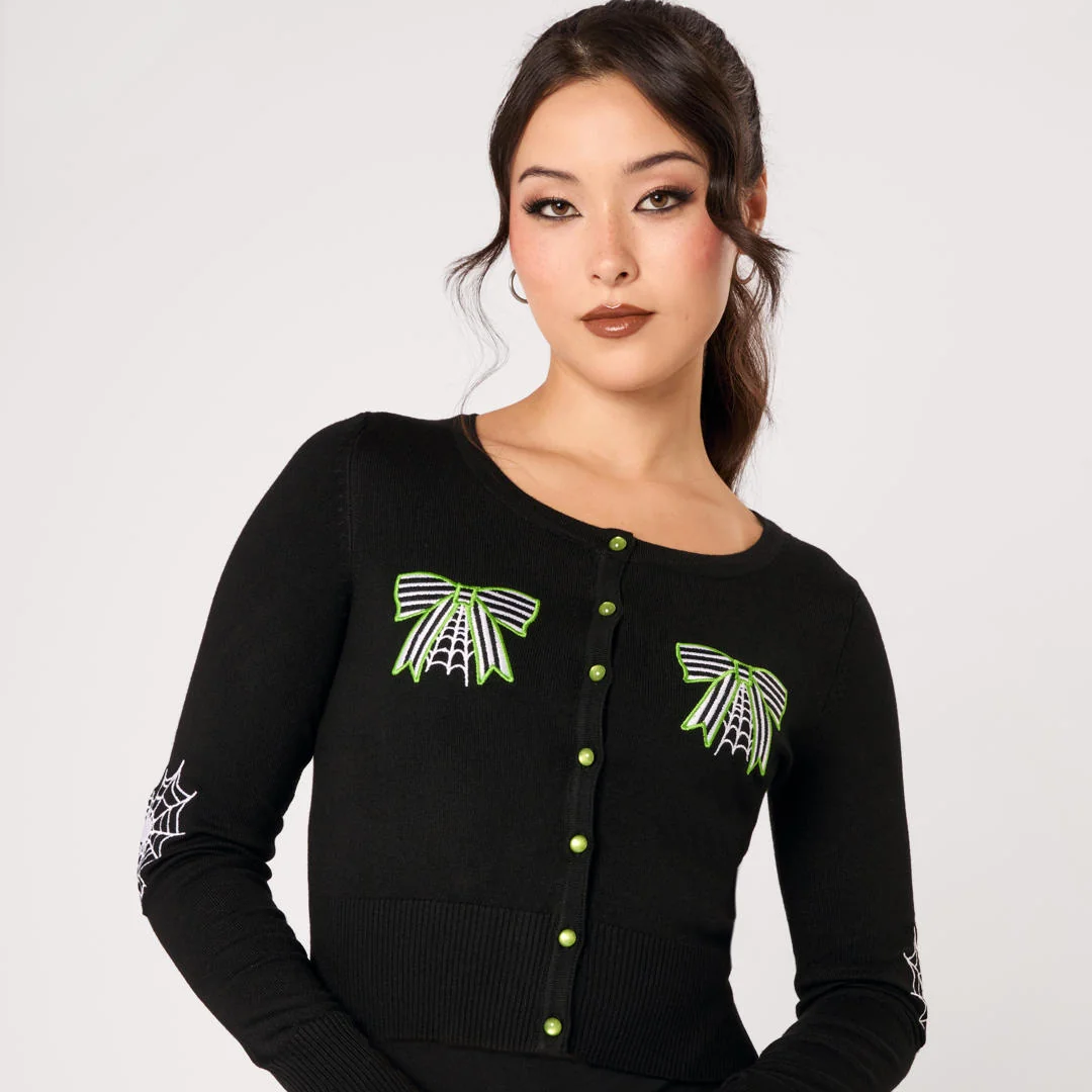 A woman modeling a black long-sleeve knit top with green and white spider-themed embroidery on the chest and sleeves, paired with black pants, against a light gray studio background.