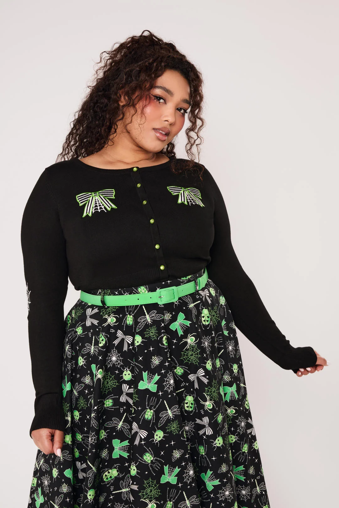 A woman modeling a black long-sleeve knit top with green and white spider-themed embroidery on the chest and sleeves, paired with black pants, against a light gray studio background.