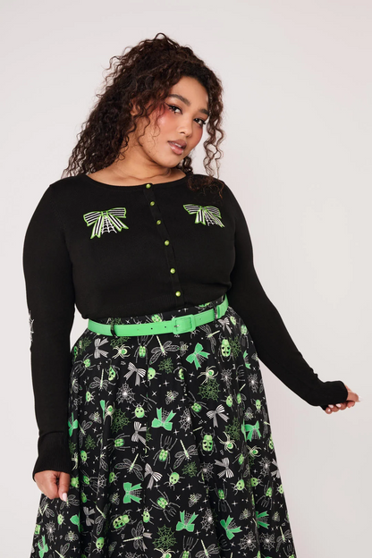 A woman modeling a black long-sleeve knit top with green and white spider-themed embroidery on the chest and sleeves, paired with black pants, against a light gray studio background.