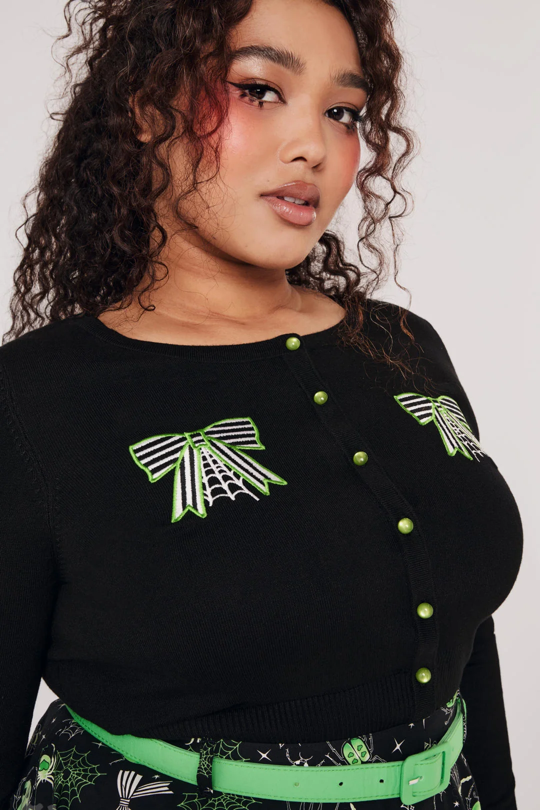 A woman modeling a black long-sleeve knit top with green and white spider-themed embroidery on the chest and sleeves, paired with black pants, against a light gray studio background.