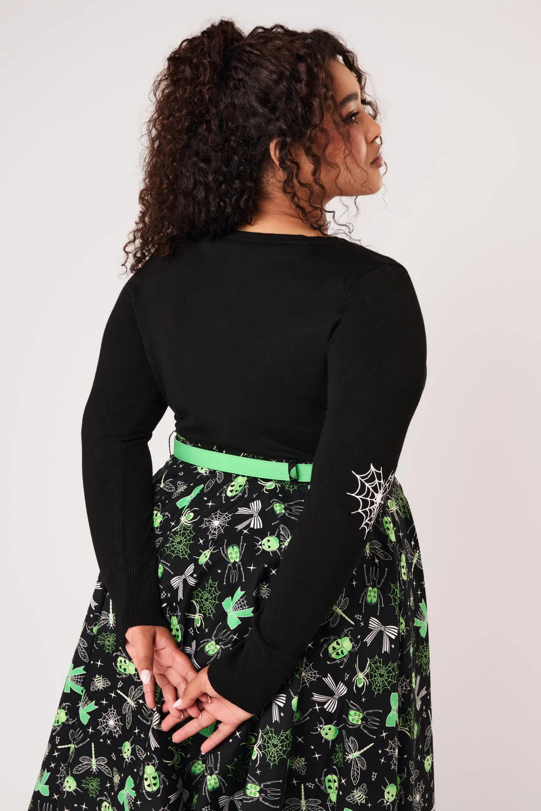 A woman modeling a black long-sleeve knit top with green and white spider-themed embroidery on the chest and sleeves, paired with black pants, against a light gray studio background.