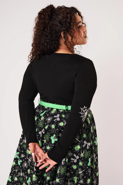 A woman modeling a black long-sleeve knit top with green and white spider-themed embroidery on the chest and sleeves, paired with black pants, against a light gray studio background.