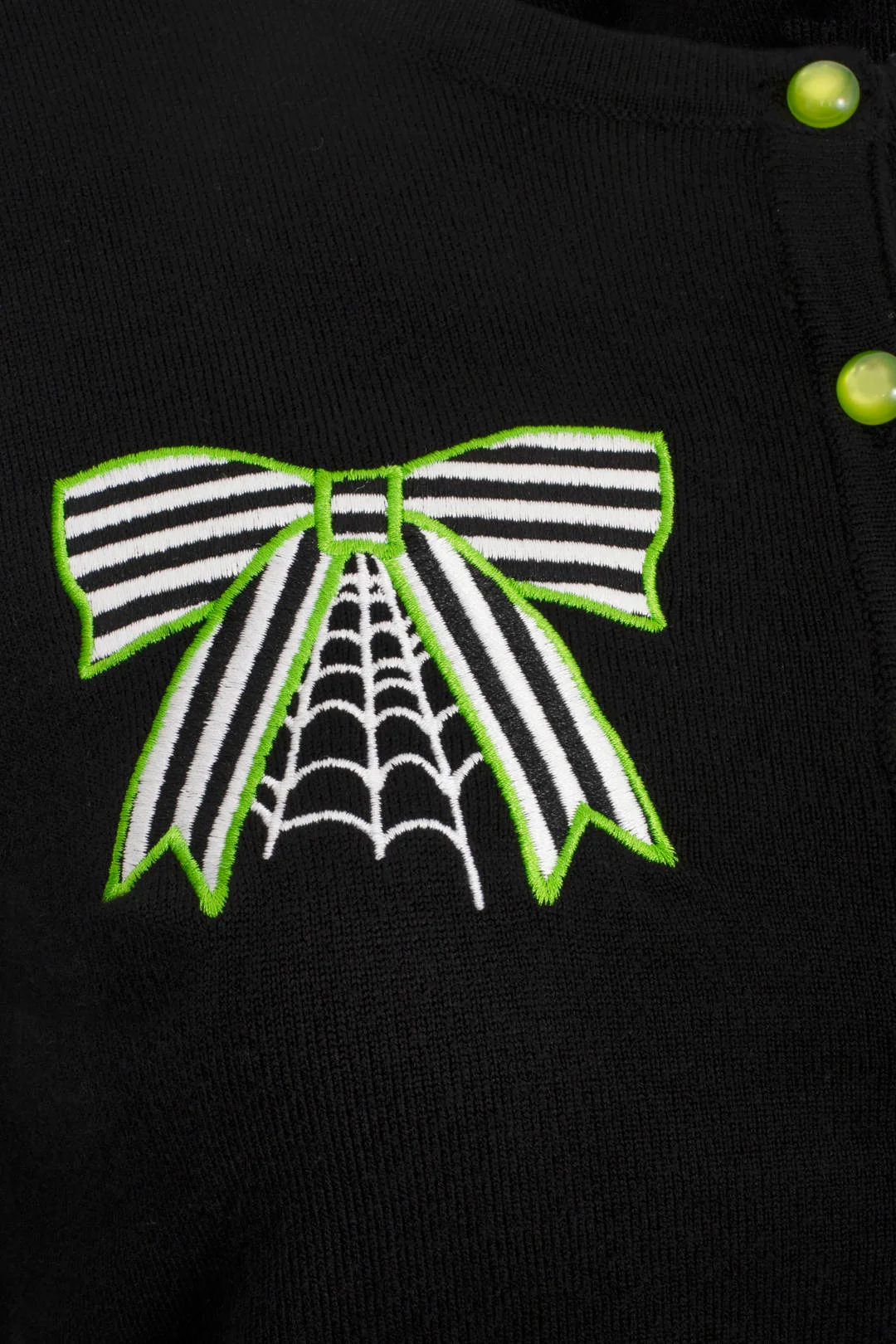 A woman modeling a black long-sleeve knit top with green and white spider-themed embroidery on the chest and sleeves, paired with black pants, against a light gray studio background.