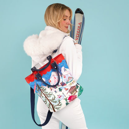 A woman in a white winter outfit with a fluffy hood carries a colorful ski-themed tote bag. She is posed against a solid light blue background, holding skis behind her back.