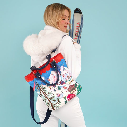 A woman in a white winter outfit with a fluffy hood carries a colorful ski-themed tote bag. She is posed against a solid light blue background, holding skis behind her back.