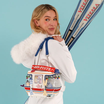 A woman in a white winter coat with a fluffy trim carries a decorative crossbody bag shaped like a snowmobile. She is holding a pair of blue and white skis against a solid light blue background.