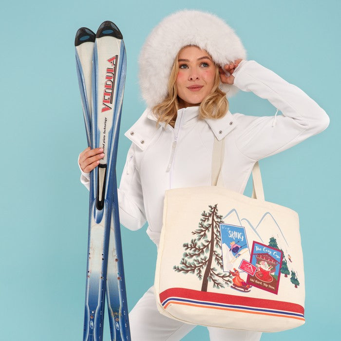 A woman in a white ski outfit and fluffy white hat holds a pair of blue skis and carries a cream canvas tote bag with a colorful ski resort graphic, posed against a light blue studio background.