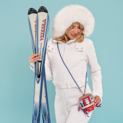 A woman in a white ski outfit with a fluffy white hat holds a pair of blue and white skis, wearing a red and white crossbody bag with a blue strap, set against a solid light blue background.