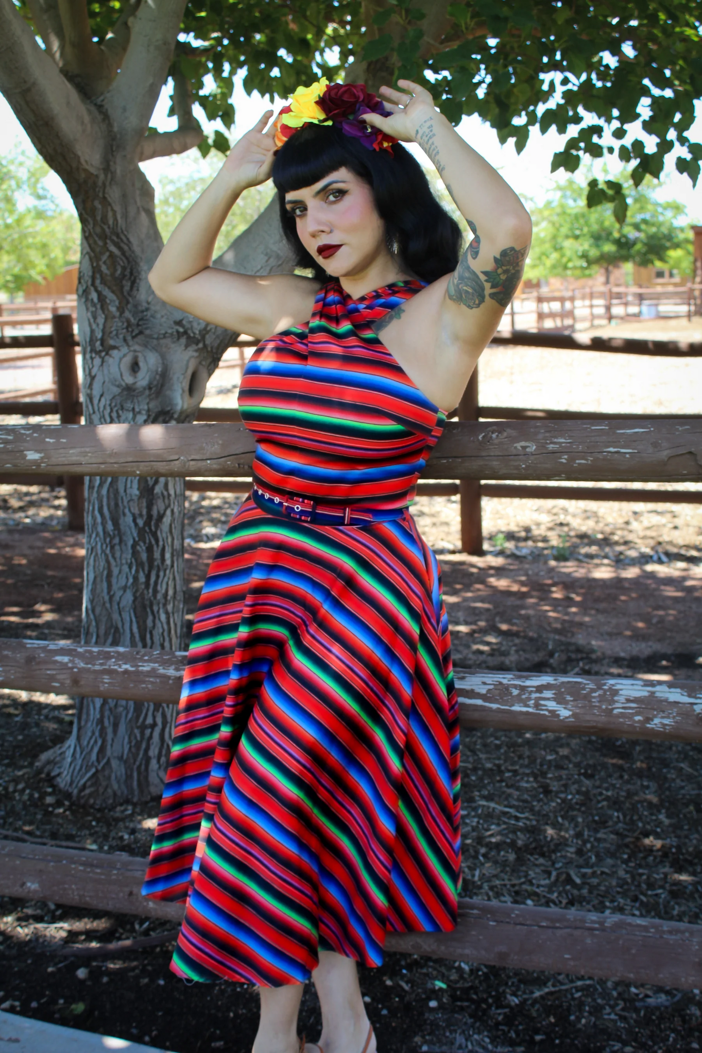 A woman posing outdoors in front of a wooden fence and tree, wearing a vibrant, multicolored horizontally striped halter dress with a matching belt and a floral headband.