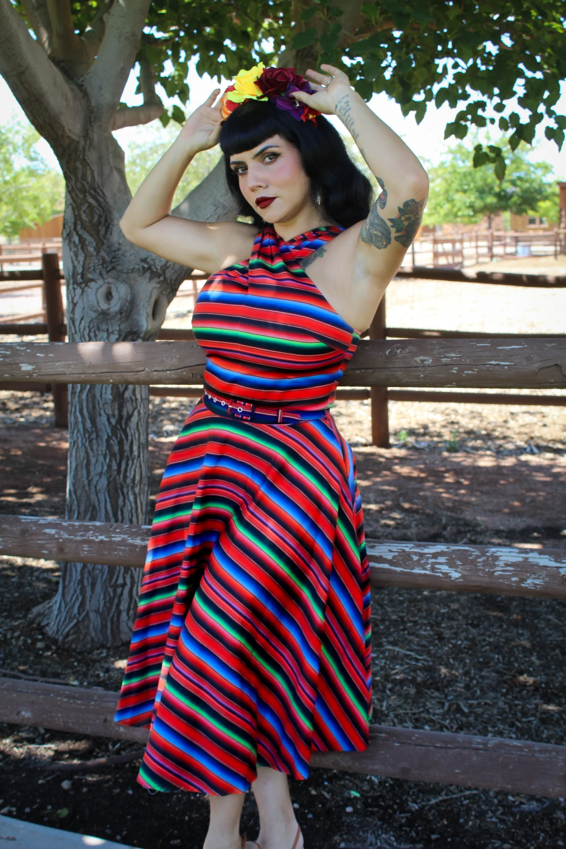 A woman posing outdoors in front of a wooden fence and tree, wearing a vibrant, multicolored horizontally striped halter dress with a matching belt and a floral headband.