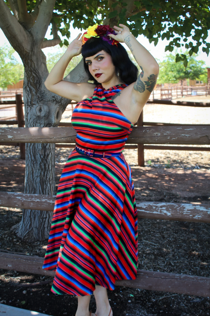 A woman posing outdoors in front of a wooden fence and tree, wearing a vibrant, multicolored horizontally striped halter dress with a matching belt and a floral headband.