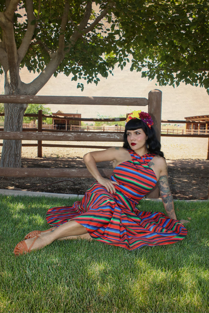 A woman wearing a colorful striped halter dress and matching headband sits on green grass in front of a wooden fence and tree.