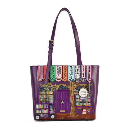 A purple leather tote bag featuring a whimsical, colorful illustration of a book-themed storefront with a tree, shelves, and books, shown against a plain white background.