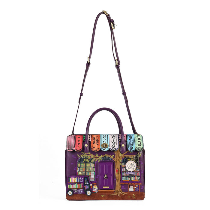 A purple leather handbag featuring a detailed illustration of a bookshop storefront on the front, with colorful book spines across the top and a long shoulder strap, shown on a plain white background.