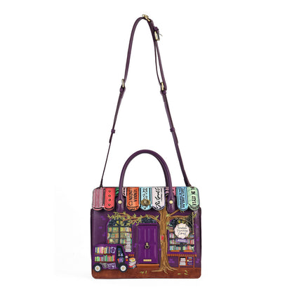 A purple leather handbag featuring a detailed illustration of a bookshop storefront on the front, with colorful book spines across the top and a long shoulder strap, shown on a plain white background.
