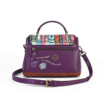 A purple leather handbag with a colorful book-patterned flap, gold-tone hardware, a top handle, and a detachable shoulder strap, photographed on a clean white background.